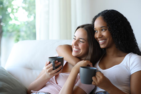 Happy interracial lesbian couple drinking coffee looking away through a window at homeの写真素材