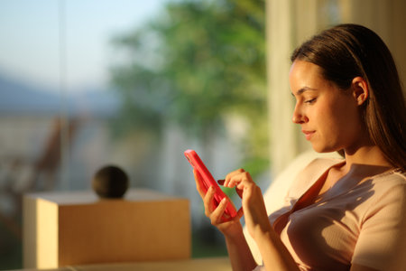 Serious woman using phone to control smart speaker at sunset at homeの写真素材