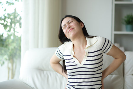 Asian woman suffering lumbago sitting on uncomfortable couch at homeの写真素材