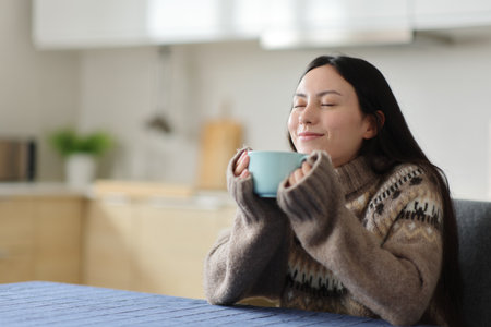 Asian woman wearing sweater in winter relaxing smelling coffee in the kitchen at homeの写真素材