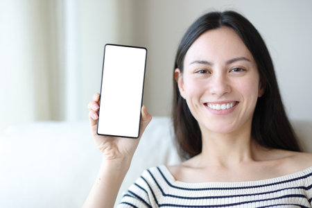 Happy asian woman showing blank phone screen sitting on a sofa at homeの写真素材