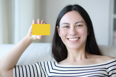 Happy asian woman showing blank credit card mock up sitting on a couch at homeの写真素材