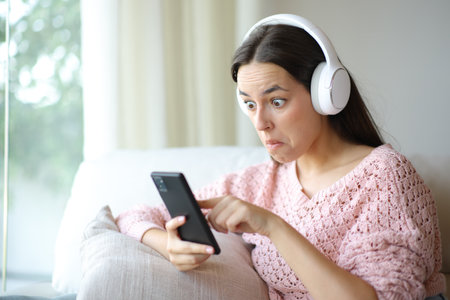 Perplexed woman listening audio with smart phone and headphone at homeの写真素材