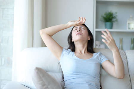 Stressed asian woman suffering heat stroke fanning with hand sitting on a sofa at homeの写真素材