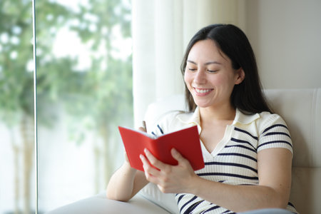 Happy asian woman writing in agenda sitting on a sofa at homeの写真素材