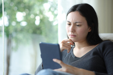 Confused asian woman reading ebook sitting on a couch at homeの写真素材