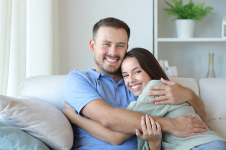 Happy couple at home hugging sitting on a couch looking at youの写真素材