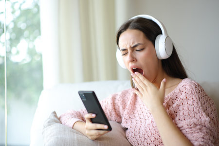 Bored woman wearing headphone yawning listening audio on phone at homeの写真素材