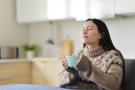 Asian woman breathing fresh air drinking coffee sitting in the kitchen in winter at homeの写真素材