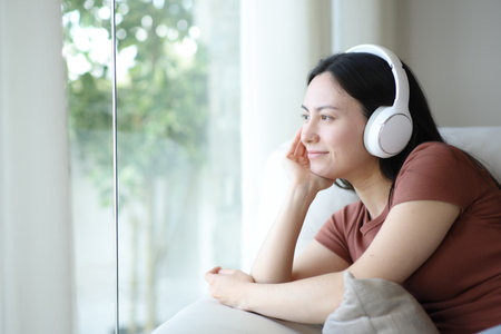 Asian woman wearing headphone relaxing listening audio looking through a window at homeの写真素材