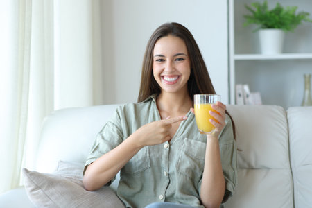 Happy woman at home holding and showing orange juice sitting on a couchの写真素材