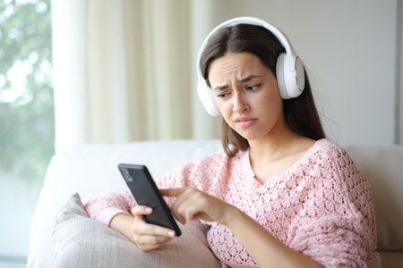 Disgusted woman checking phone using headphone at homeの写真素材