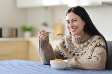 Happy asian woman in winter eating cereals looking at you in the kitchen at homeの写真素材