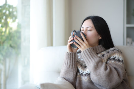 Asian woman in winter drinking coffee or tea sitting on a couch at homeの写真素材