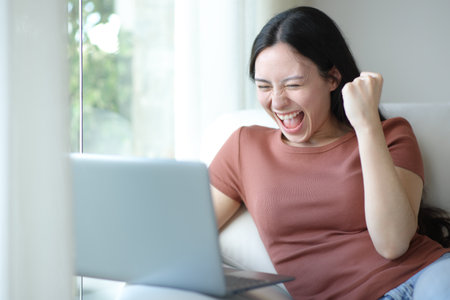 Excited woman using laptop sitting on a couch at homeの写真素材