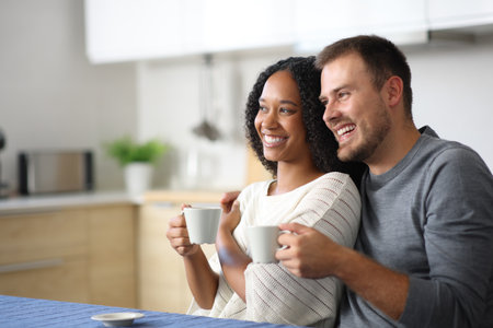 Happy interracial couple drinking coffee in the kitchen looking away at homeの写真素材
