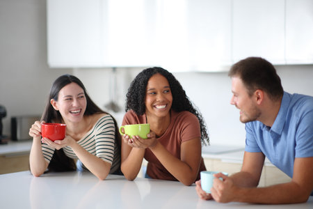 Three interracial friends talking and drinking coffee or tea in the kitchen at homeの写真素材