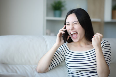 Excited asian woman talking on phone sitting on a couch at homeの写真素材