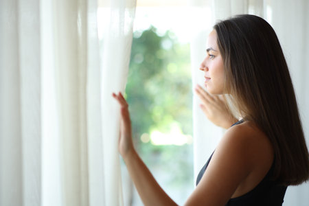 Serious woman looking through a window opening curtain at home or hotel roomの写真素材