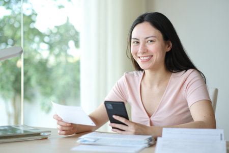 Asian woman holding bill and phone posing looking at you at homeの写真素材