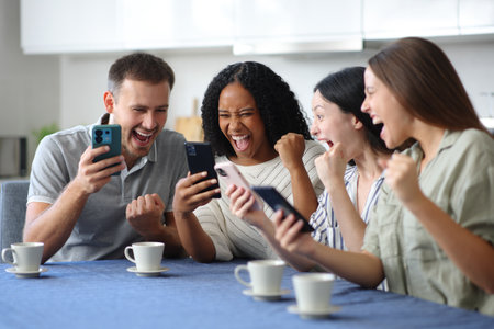 Excited friends celebrating checking their phones in the kitchen at homeの写真素材