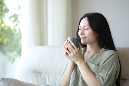 Satisfied asian woman enjoying smelling coffee sitting on a couch at homeの写真素材