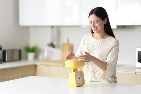 Happy asian woman weighing food with scale in the kitchen at homeの写真素材