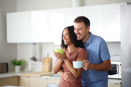 Happy interracial couple drinking and looking away in the kitchen at homeの写真素材