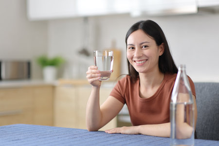 Happy asian woman holding glass of bottled water in the kitchen at homeの写真素材