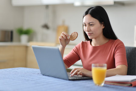 Asian student e-learning using laptop and eating cookie at breakfast in the kitchen at homeの写真素材
