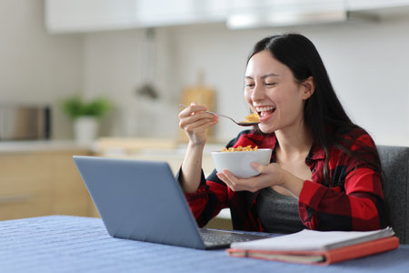 Happy asian student e-learning with a laptop eating cereals for breakfast in the kitchen at homeの写真素材