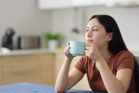 Distracted asian woman drinking coffee in the kitchen at homeの写真素材