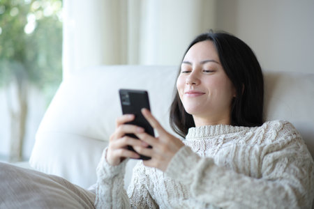 Satisfied asian woman checking cell phone sitting on a sofa at homeの写真素材