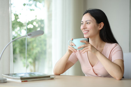 Happy asian woman drinking tea looking through a window in a room at homeの写真素材