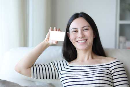 Happy asian woman showing blank card sitting on a couch at homeの写真素材