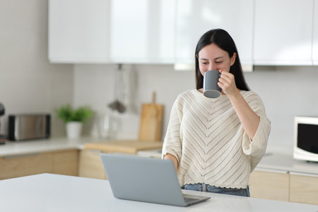 Asian woman drinking coffee and checking laptop standing in the kitchen at homeの写真素材