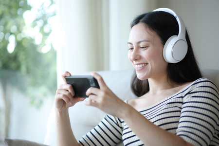 Happy asian woman wearing headphone watching videos on phone sitting on a sofa at home