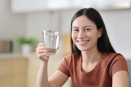 Happy asian woman holding water glass looking at camera in the kitchen at homeの写真素材