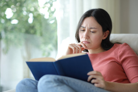 Sad asian woman reading a paper book sitting on a couch at homeの写真素材