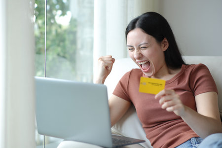 Excited asian woman buying online with laptop and credit card sitting at homeの写真素材