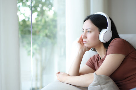 Asian woman wearing headphone listening music looking through a window at homeの写真素材