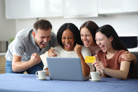 Four interracial excited friends paying online with laptop and credit card celebrating in the kitchen at homeの写真素材