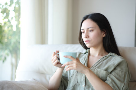 Apathetic asian woman drinking and complaining sitting on a sofa at homeの写真素材