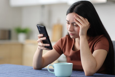 Worried asian woman checking smart phone complaining in the kitchen at homeの写真素材