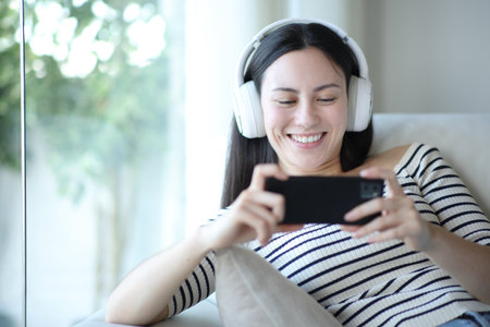 Happy asian woman watching videos on phone sitting on a couch at homeの写真素材