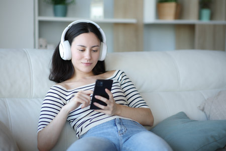 Asian woman watching media content on phone sitting on a couch at homeの写真素材