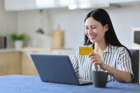 Happy asian woman paying online with laptop and credit card in the kitchenの写真素材