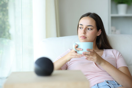 Woman relaxing drinking coffee with smart speaker in foreground at homeの写真素材