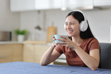 Happy asian woman wearing headphone listening audio and drinking in the kitchen at homeの写真素材