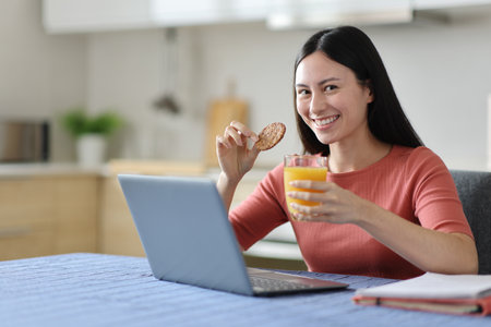 Happy asian student e-learning posing at breakfast with a laptop holding cookie and orange juice in the kitchen at homeの写真素材